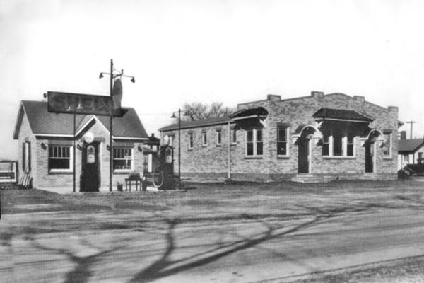black and white photo small gable roof brick building with chimney and cross gable over central door, windows on each side, 2 gas pumps and Shell signs. To the right a brick buidling two doors, five windows with awnings