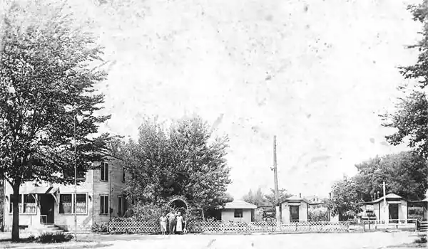 Black-white 1940s. Left: house picket fence with archway and people. Cabins behind