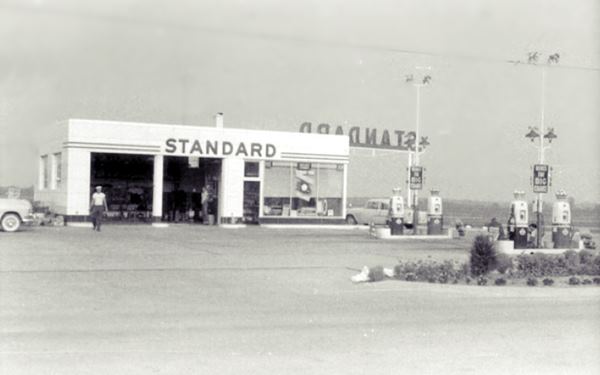 black and white 1950s photo of an oblong-box Standard Service station, 4 gas pumps, 2 cars and a man by the two service bays