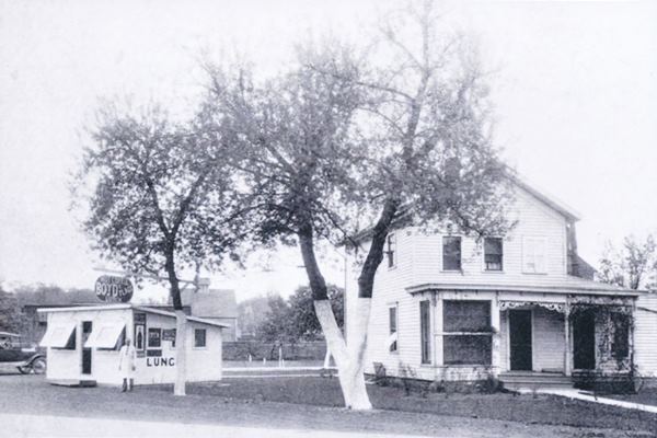 black and white 1930s, two men by a single story small lunch diner, with signs. Trees, lawn and 2 story woodframe gable roofed home, railroad behind