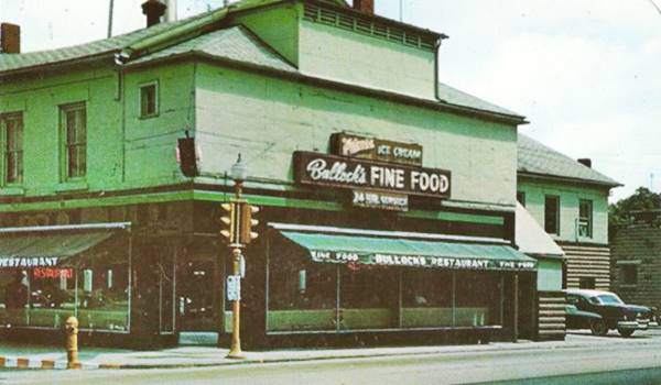 green building 2 stories, on a corner sign reads BULLOCKS FINE FOODS RESTAURANT, US 66 runs in front, odd hip roof, car parked right side