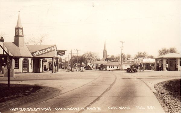 black and white 1937, several gas stations with some cars at a paved intersection in a town, church bhind a hip roofed cafe