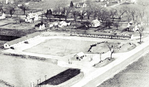 black and white 1950s aerial view: L-layout motel to the right and gas station to the bottom left. US 24 runs on the right