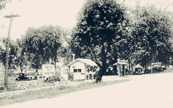 black and white 1933 US 66 in the foreground, park with trees, building, cars parked and sign reading CHENOAS FREE TOURIST PARK