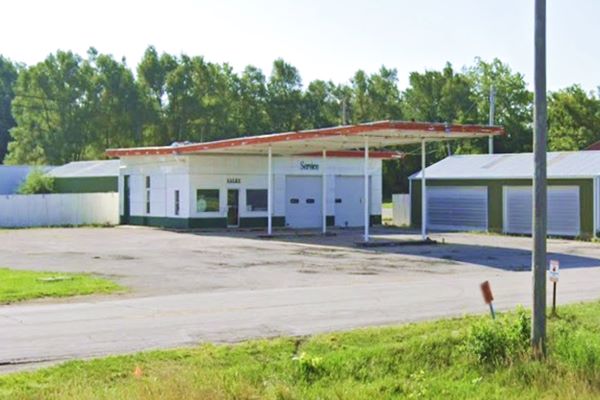 icebox shaped former gas station, highway in front, 2 pump islands under flat canopy, trees behind it