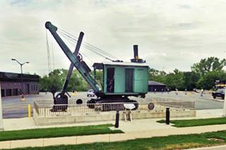 green early 1900s steam powered bucket shovel, low fence around it, US 66 shield on fence, seen from Route 66