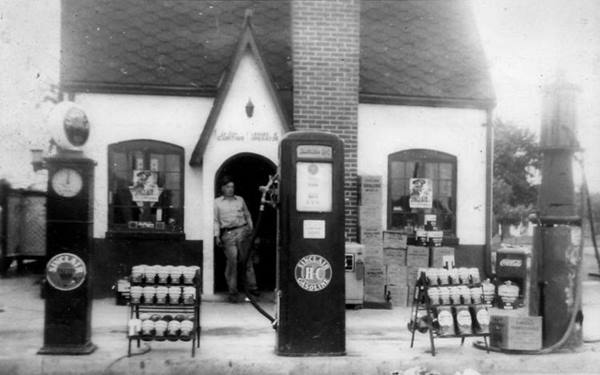 black and white 1920s cottage shaped Sinclair gas station, 3 pumps, man by door