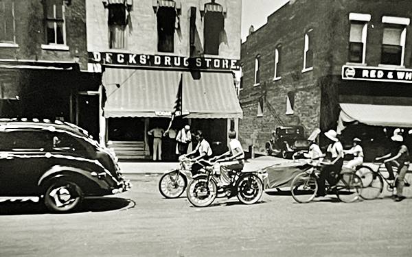 black and white vintage 1940s photo of brick building with sign "Deck’s Drugstore" over entrance, children on bikes ride along the street and a 1940s car
