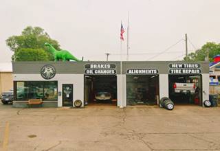 box shaped 2 bay, black garage tire ship, with flat roof on which green dinosaur stands, seen from Route 66