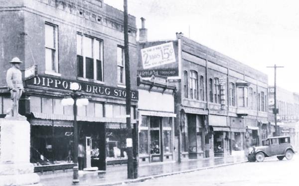 black and white 1920s photo looking north along Macoupin St: A drugstore and Doughboy statue to the left, cars parked and brick fronted stores face the street