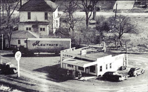 black and white, c.1950s Texaco gas station and cafe on a curved corner, parking area, truck semi behind. 2 story hip roof home behind