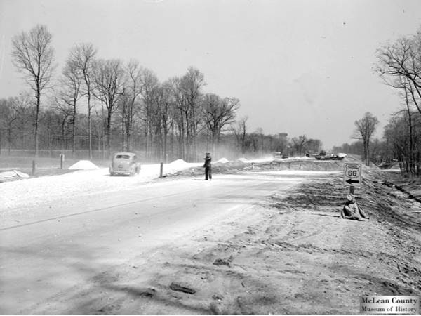 US66 shield, man guiding traffic at a road construction site. Trees line the highway. Old road forks to the right. Black and white 1946