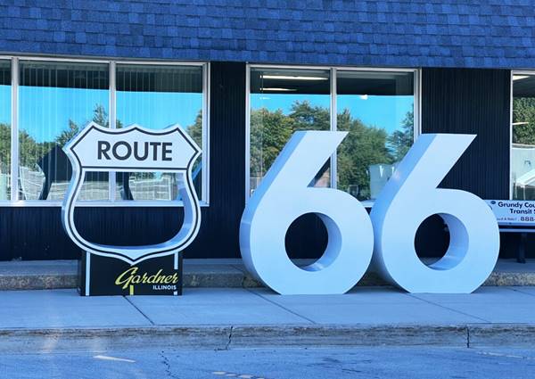 large sized hollow US66 shield on pedestal that reads GARDNER and two sixes on a sidewalk, white colored. A monument