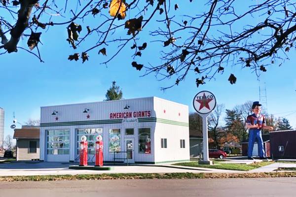 color, box-shaped Texaco signed recreated gas station, gas pumps and a giant muffler man (right) street in front, buildings beyond, leaves above