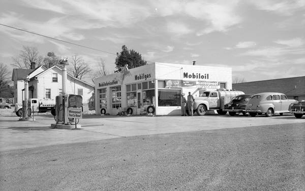 black and white 1951: icebox shaped Mobil gas station, cars, 2 men by a tanker truck 4 pumps, glazed corner office by men, 2 service bays
