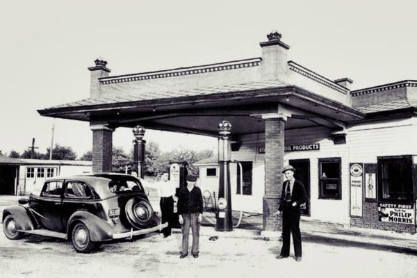 black and white c.1930s Standard Oil proeducts gas station, flat canopy pent roof around it and office, brick columns, 3 pumps, a car. 3 men and a woman stand by it