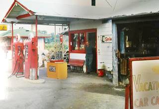 Groll’s gas c.1950s Dwight color, 1950s, 2 pumps under gable roof on a woodframe building, man at office door