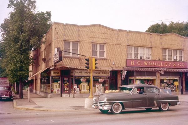 yellow brick corner building, a pharmacy c.1950s, cars, people, signs, color.