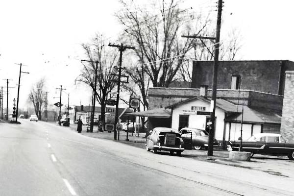 black and white 1950s photo of a highway with cars, traction rail tracks, building to the right, and a gas station beyond