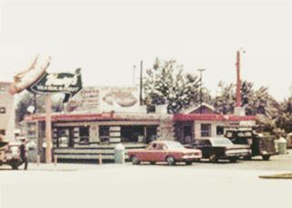 Henry’s hot dog drive in in the 1960s color, 1960s, hot dog shaped neon sign atop a box shaped hot dog stand, cars around it