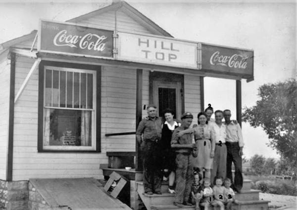 black and white 1940s, several adults and childrens on entry steps, gable roof, single story, sign over doorway reads HILL TOP Coca Cola 
