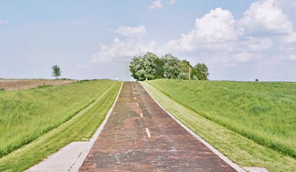 Brick paved Route 66 on a hill, fields on both sides, white concrete curb and trees next to highway on the hilltop