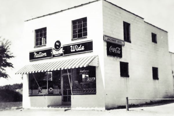black and white photo c1940s of a white, 2-story, block-shaped store with signs over entrance (display windows on each side of the door) flat roof