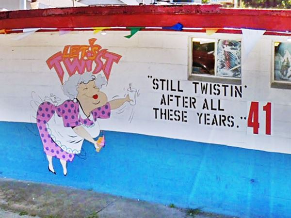 cartoon of a mature woman in polka dot dress with an apron holding an ice cream in one hand and a paper cup in the other words around her say "Still Twistin’ after all these years 41"