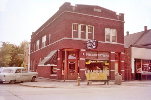 red brick corner building, a pharmacy c.1950s, cars, people, signs, color.