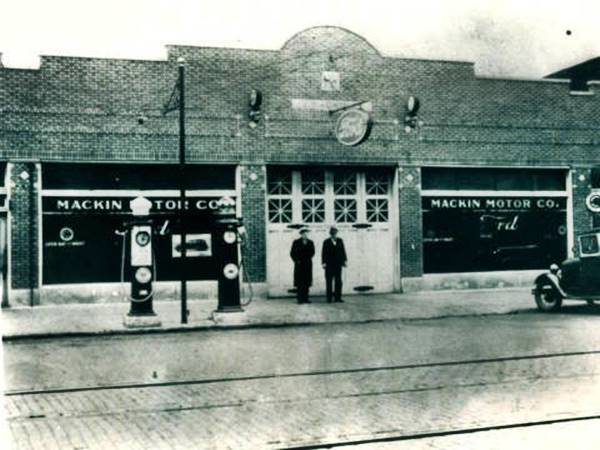 black and white c.1928, two men by brick building, garage door, large windows on both sides with MACKIN MOTOR CO written on them, 2 gas pumps left, car to the right, street in front