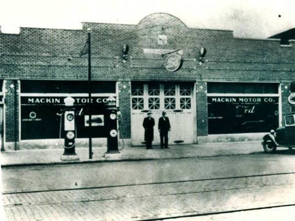 Mackin Motor Co c.1928 Lockport IL black and white c.1928, two men by brick building, garage door, large windows on both sides with MACKIN MOTOR CO written on them, 2 gas pumps left, car to the right, street in front