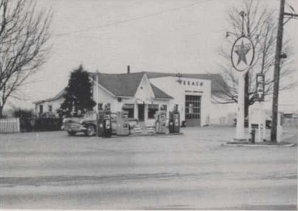 black and white 1958 gable roof cafe (left) Texaco sign, pumps and car, box shaped garge single bay (right)