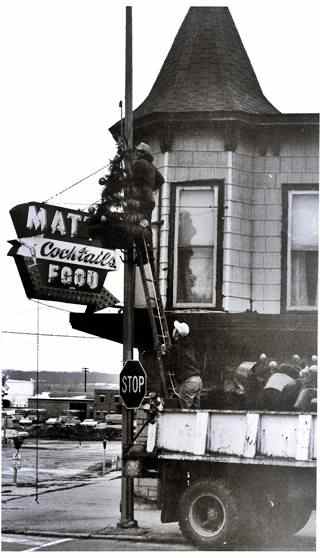 Matt’s Inn, 1974, Lemont IL black and white, man on a ladder resting in truck, removing Xmas decoration from light post by 2 story building with neon sign reading MATT COCKTAILS FOOD