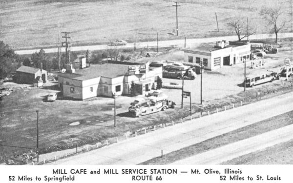 black and white 1950s postcard four lane highway in foreground, two buildings with parked cars, trees, cafe on the left, gas station on the right, highway behind the premises