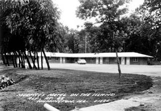 black and white, gable roof 1 floor motel surrounded by trees, 1950s car parked by it