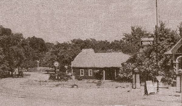 sepia, 1937, road curves, a garage to the left, and a canopy and STANDARD SERVICE sign to the right, trees beyond