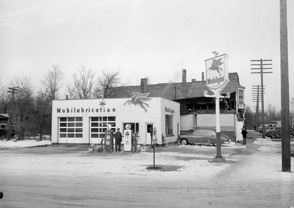 1950 black and white, box shaped Mobil station, 2 gas pumps, attendant, car parked