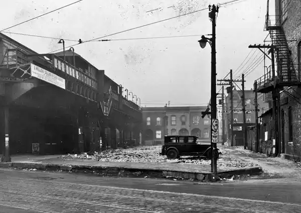 black and white. Ogden and elevated rail station, garbage strewn on parking area, car by a light post with shields of US 66, US 32, IL 4 and IL 18