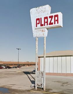 faded white 1970s sign with red letters spelling: PLAZA, in a vacant parking lot