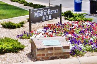 marker set in red paving bricks by sidewalk, museum sign next to it, and flower bed