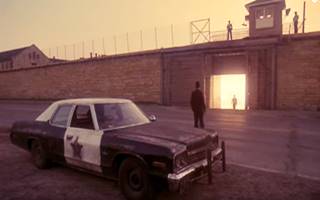 Prison release scene from THE BLUES BROTHERS movie man walks from open prision gate, lit from behind to a car where another man waits. Tall walls, guards on them