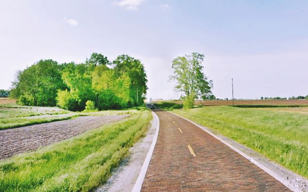 Brick paved Route 66 on a hill curves by a stream, fields on both sides, some trees to the left, white concrete curb