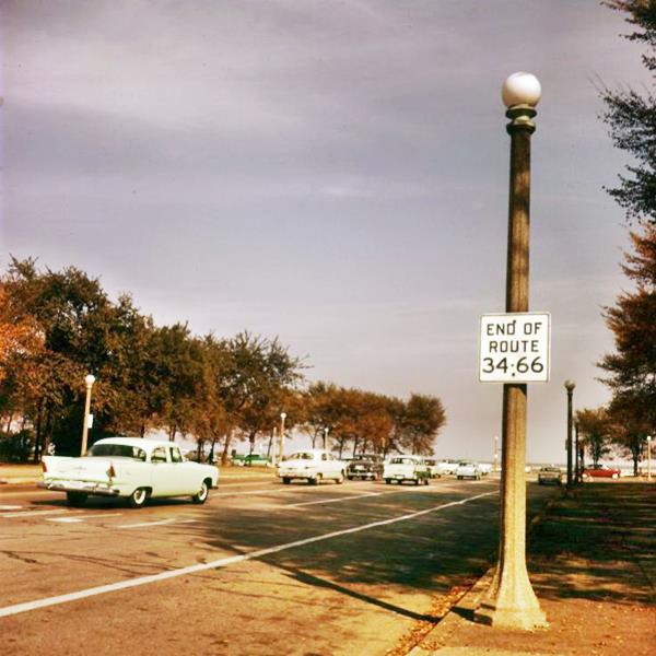cars next to End of US 66 and 34 sign in the park in a 1950s color photo