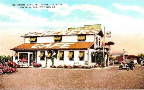 1930s colorized postcard with gable roof two story building, words "Scherer’s Cafe" written on roof. Behind is a gas station and a car