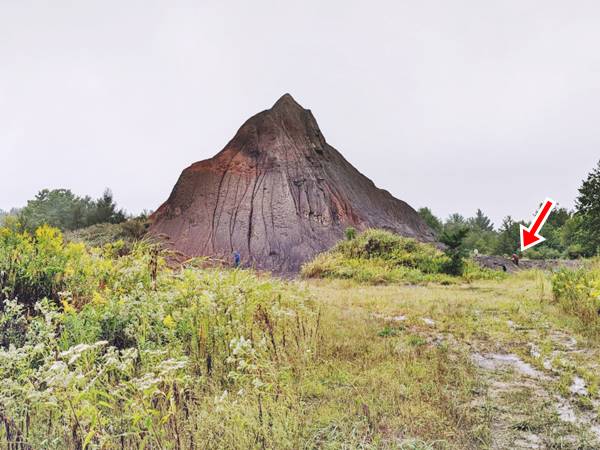 gray pile of slag 100 ft tall in an area with trees and bushes, red arrow marks a man standing by it