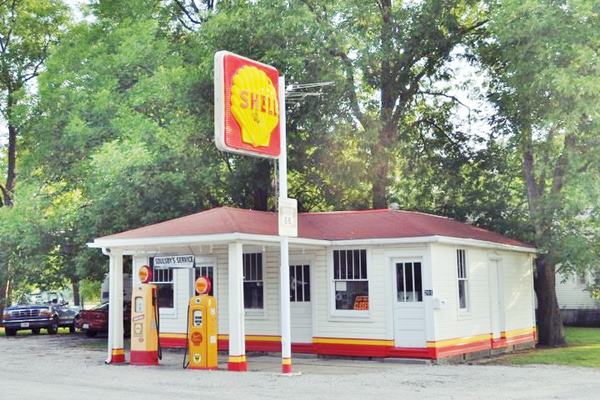vintage red hipped roof and canopy, white woodframe 1920s gas station with pumps and Shell signage, neatly restored, trees behind it, US66 in front.