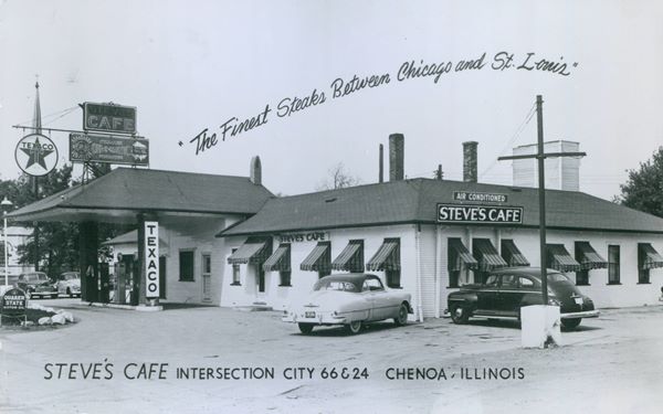 black and white 1941: hip roof building, a cafe, with signs saying STEVES CAFE, and Texaco pumps and signs under a hipped canopy. Church Steeple behind, cars
