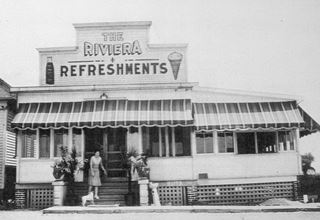 black and white 1930s woman with dog by woodframe building entrance steps sign above her reads THE RIVIERA REFRESHMENTS