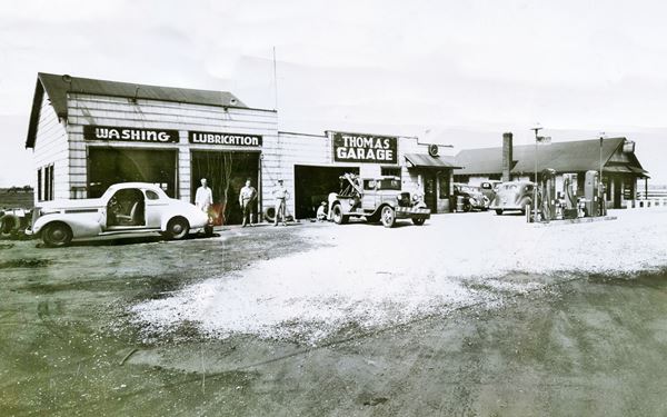 black and white picture c.1940s of two buildings one with service bays, gas pumps, and cars 