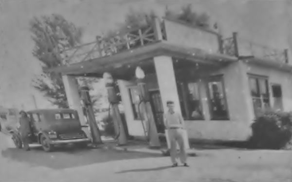 black and white 1936. Man by gas station, gas pumps, canopy and woman by car